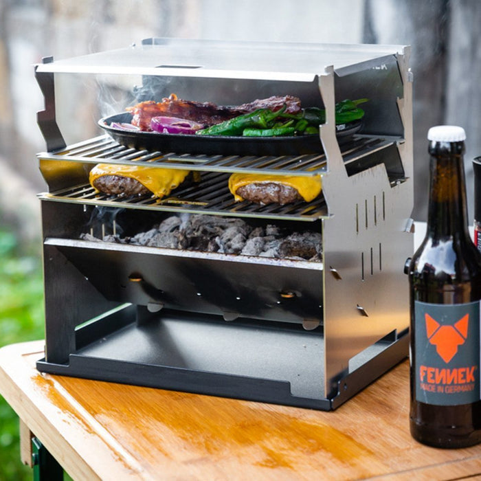 Fennek Grill im Einsatz mit zweitem Grillrost, unten ein Grillrost zum Grillen von Cheeseburger und oben ein Grillrost zum Warmhalten. Daneben zwei Flaschen mit FENNEK Label.