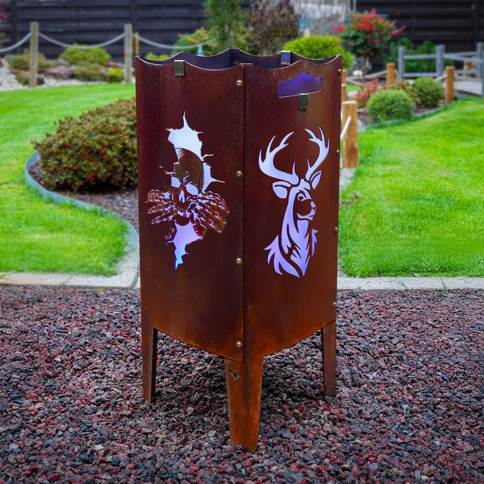 Die FENNEK Feuersäule aus Stahl mit Totenkopf und Hirsch Motiv steht auf einem Kiesbett in einem schönem Garten. Die Feuersäule hat einen charmanten Rostlook und leuchtet, dank des LED Lichtersets lila.