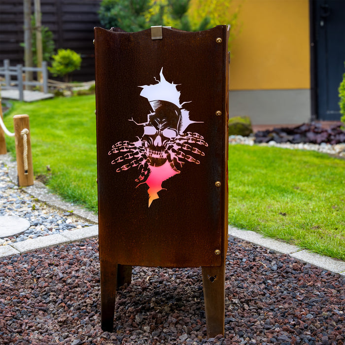 Die FENNEK Feuersäule aus Stahl mit Totenkopf steht auf einem Kiesbett in einem schönem Garten. Die Feuersäule hat einen charmanten Rostlook und leuchtet, dank des LED Lichtersets orange.