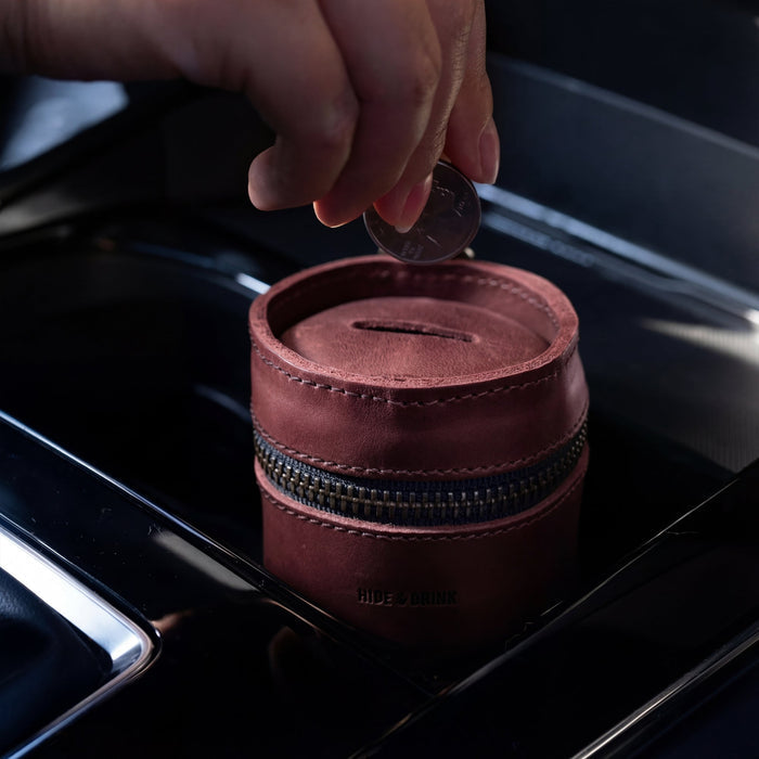 A person placing a coin into Hide & Drink's Bucket Coin Release, perfectly fitting within a car's console. This image showcases how the handy cash organizer, crafted from full grain leather, serves as an ideal money box and zippered pouch for everyday use.