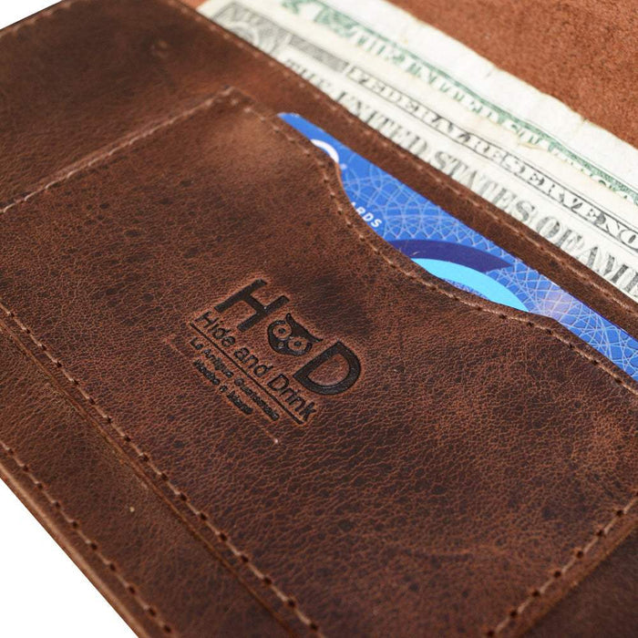 Close-up of the logo on the Brown Double Snap Folio Wallet by Hide & Drink, emphasizing its quality craftsmanship. Made from full grain leather, this rectangular flat bill holder with card slot serves as a night bag and travel accessory. Hand sewn by artisans in La Antigua Guatemala & Oaxaca Mexico, it's a testament to small batch production.