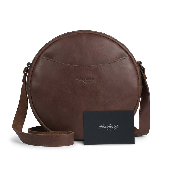Heather's Female Rounded Crossbody Hand Bag with a note peeking out, depicting its practical use. Crafted from Full Grain Leather, this zippered handbag for women and shoulder bag for girls is both stylish and functional. Handmade in La Antigua Guatemala, showcasing the artisan's hand cut, hand sewn, and small batch production techniques.