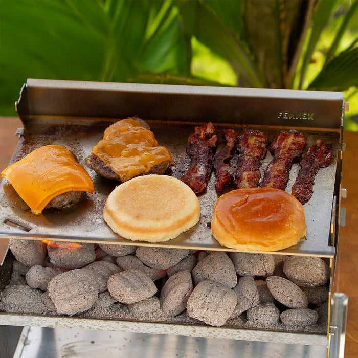 Zubereitung von Cheeseburger und Bacon auf einer Edelstahl Planchaplatte die auf dem Holzkohlegrill aufliegt und mit Holzkohlebriketts betrieben wird.