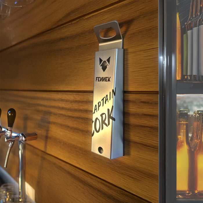 Der FENNEK Flaschenöffner Captain Cork mit Magnet ist an einer Holzverkleideten Wand einer Bar angeklebt. Rechts davon ist ein voller Kühlschrank zu sehen. Links kann man Zapfhähne sehen.
