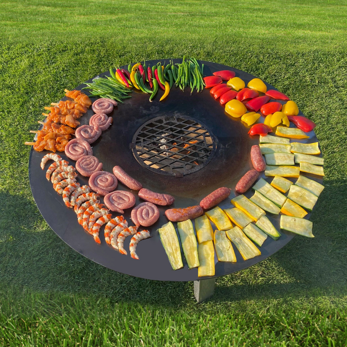 Szenebild FENNEK Feuerplatte - 80 cm inkl. optionalem Zubehör Grillrost. Auf der Feuerplatte werden Würste, Gemüse und diverse Paprika gegrillt.