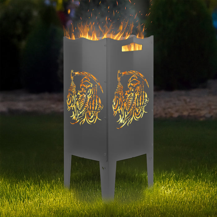 FENNEK Feuersäule Stahl mit dem Motiv Sensenmann - in der Feuersäule brennt ein Feuer, man sieht das Motiv sehr schön leuchten in der Abenddämmerung in einem Garten. Die Feuersäule steht auf Gras.