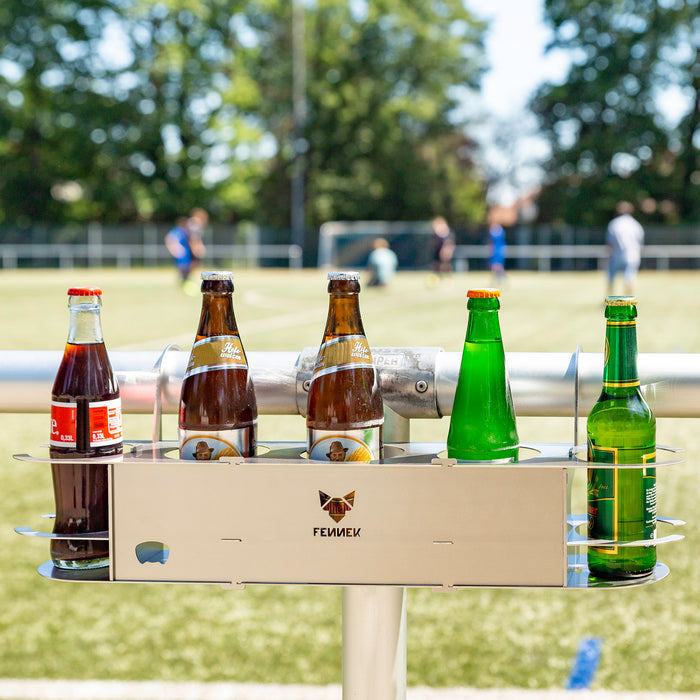 Fünf Glasflaschen in einem Getränkehalter aus Edelstahl mit FENNEK-Logo in der Mitte, befestigt am Fussballspielfeldrand. Im Hintergrund unscharf Fussballspieler.