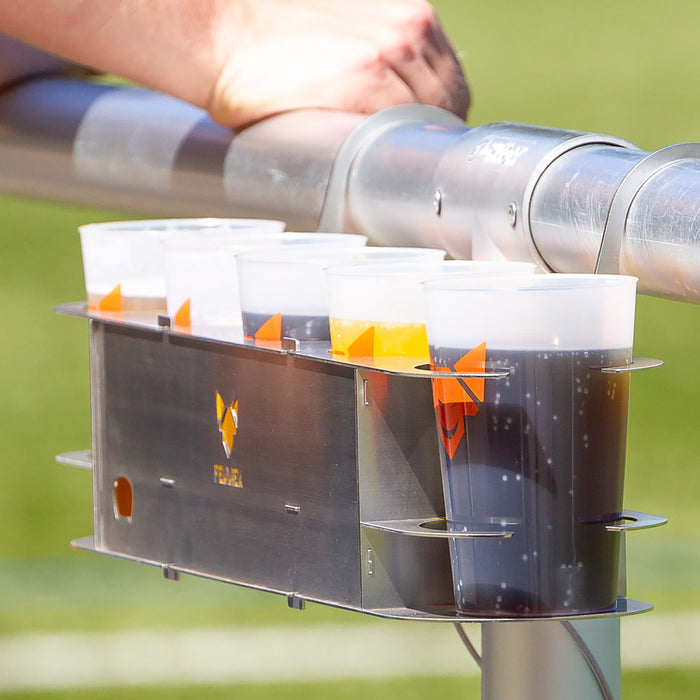 Fünf gefüllte Getränkebecher in einem Getränkehalter aus Edelstahl mit FENNEK-Logo in der Mitte, befestigt am Fussballspielfeldrand.