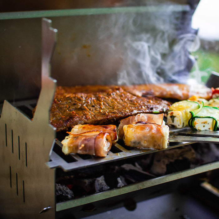 Bild zeigt den FENNEK Grill in Betrieb.  Auf dem Grillrost brutzelt unter anderem Gemüse und Fleisch vor sich hin. Außerdem ist der dazugehörige Rostheber eingehakt. Dieser kann aber auch als Flaschenöffner genutzt werden.