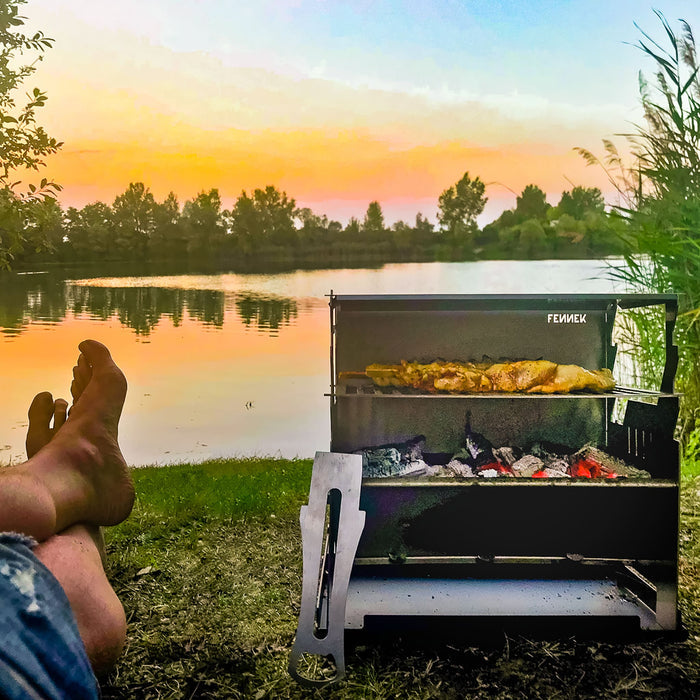 Fennek Grill mit glühender Kohle und Grillfleisch auf dem Rost bei Sonnenuntergang am See.