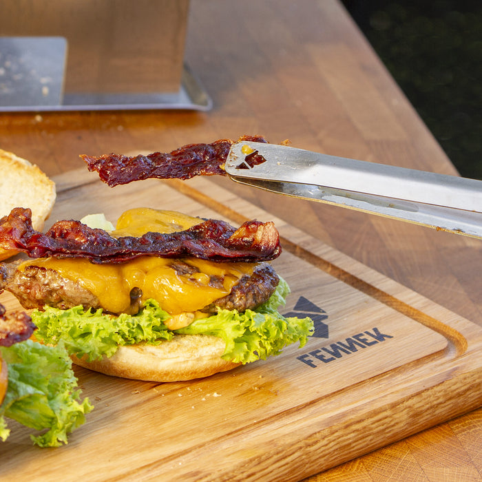 Mit Grillzange von Fennek wird ein Stück knusprig gebratener Bacon auf einen Hamburger gelegt.