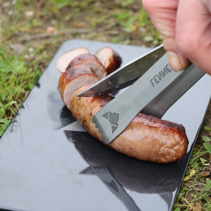 Kleine Grillzange aus Titan mit laser-graviertem FENNEK-Logo und Titanium-Schriftzug im Einsatz beim Schneiden einer Bratwurst