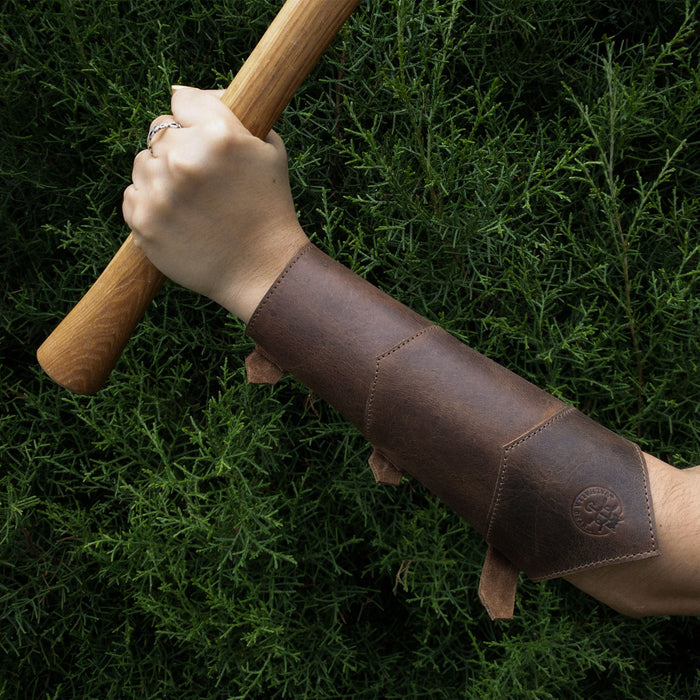 A powerful grip on a wooden staff, showing the Valhalla Gear Forearm Protector for Bow Shooting Practice worn as a Female Archery Bracer. This sturdy armband, designed with adjustable buckle straps, emphasizes the characteristics of high-quality full grain leather, perfect for cosplay and outdoor sports, along with its handmade origins from artisan craftsmen in La Antigua Guatemala.