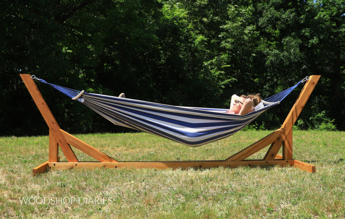 Person relaxing in a striped hammock on a wooden stand with greenery in the background