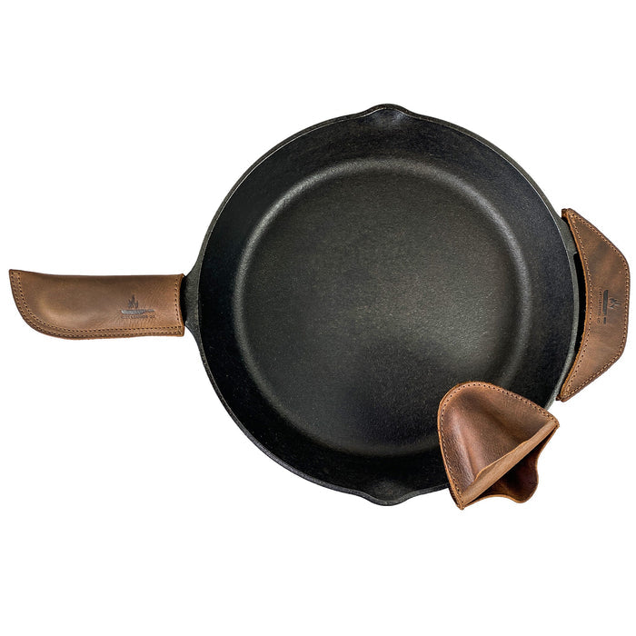 This image highlights the Hot Handle Holders Set of 3 by Cast Leather Co., elegantly displaying a cast iron skillet complemented by full grain leather grip holders. Designed as a panhandle, side kick, and grip mitt, they provide reliable assist grips for stately cast iron skillets and pans, showcasing artisan craftsmanship from La Antigua Guatemala & Oaxaca Mexico through small batch production and hand finishing.