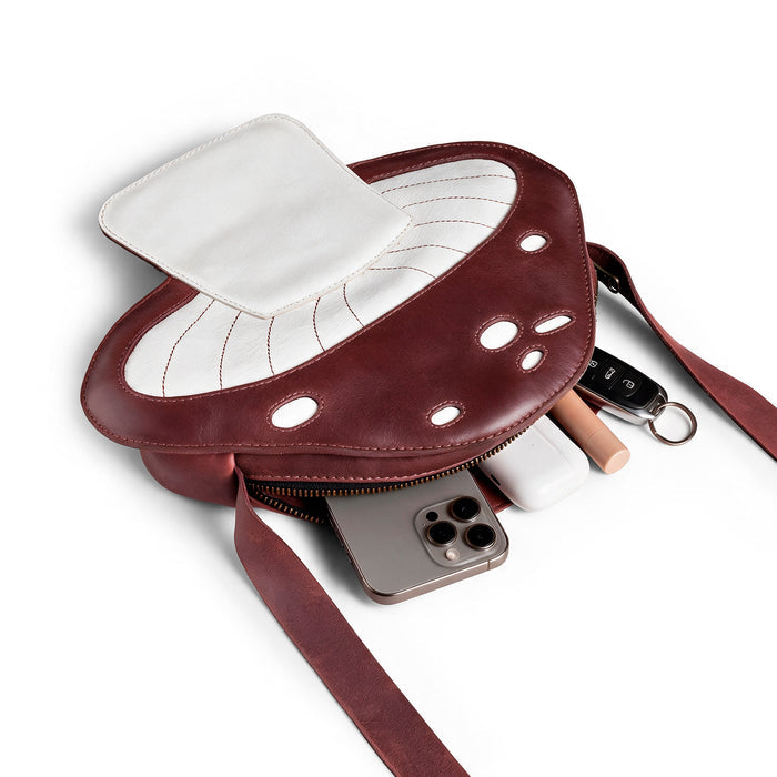 The Mushroom-Shaped Leather Shoulder Bag by Tudi Frudi is shown here filled with everyday items, demonstrating its practicality as a crossbody bag. This playful bag, made from Full Grain Leather, serves well as a travel or messenger bag, reflecting the quality of small batch production with artisan craftsmanship from La Antigua Guatemala.