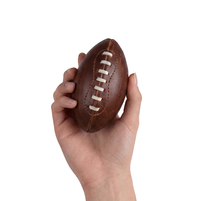 A hand holding a Mini American Football by Hide & Drink, demonstrating its small size and fine craftsmanship. This hand cut ball, made from full grain leather, makes for an excellent keepsake or display item, showcasing the brand's dedication to artisanal production.
