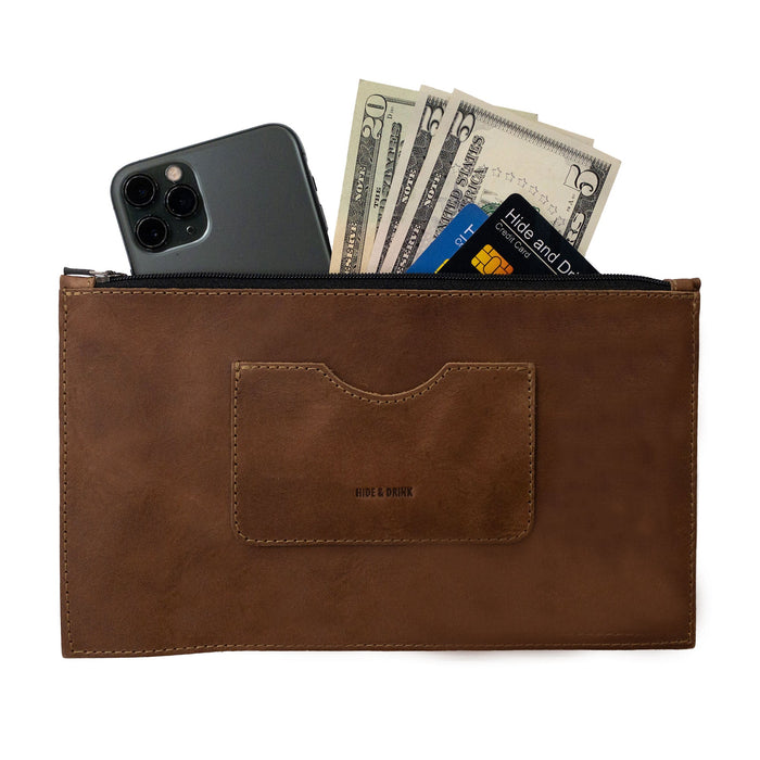 A brown rectangular zippered handbag by Hide & Drink featuring cash and a smartphone inside, designed for women. This full grain leather purse comes with a card slot, reflecting artisan craftsmanship and small batch production, hand cut and sewn in La Antigua Guatemala & Oaxaca Mexico.