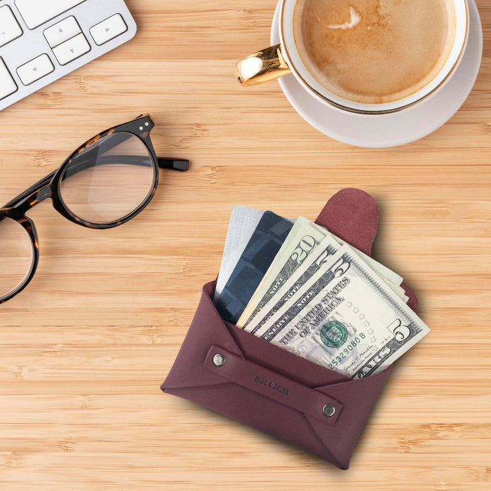 Hide & Drink's Snapless Business Card Holder on a desk with coffee. This chic wallet serves as a versatile Snapless Business Card Holder for Men and Women, providing a sturdy pouch and money organizer. Expertly made of Full Grain Leather, this product showcases small batch production with hand cut, hand sewn, and fire branded details from artisans in La Antigua Guatemala & Oaxaca Mexico.