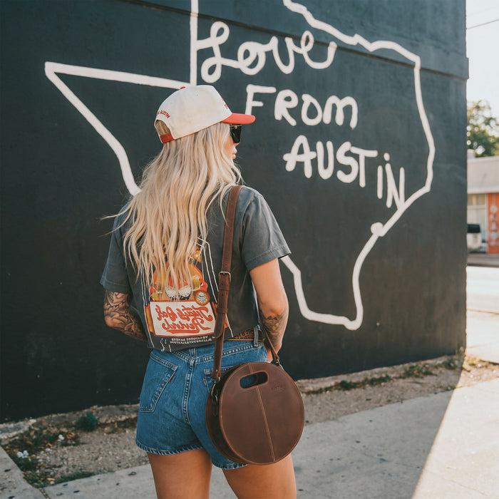 The Pecan Shoulder Bag by Austin Genuine, worn casually against a mural in Austin, Texas, exemplifying a unique blend of style and practicality, crafted from full grain leather with a focus on small batch production, hand cut and hand sewn, showcasing artisan work from La Antigua Guatemala & Oaxaca Mexico.