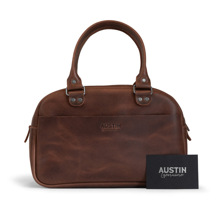 The Rainey Handbag by Austin Genuine displayed alongside an artisan card, emphasizing its quality and heritage. Constructed from Full Grain Leather, this stylish piece represents Small Batch Production, showcasing Hand Cut, Hand Sewn craftsmanship and a Fire Branded logo, made by artisans in La Antigua Guatemala & Oaxaca Mexico.