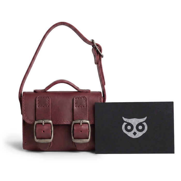 The burgundy Tiny Messenger Bag by Hide & Drink, featured alongside its tags. This Mini Messenger Bag Charm in Full Grain Leather serves as an artisan keychain or handbag accessory, handmade with care in La Antigua Guatemala.