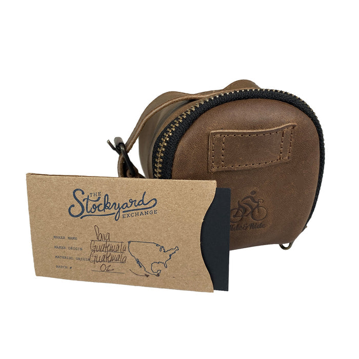 Hide & Ride Under Seat Pouch displayed alongside a special card showcasing its artisan craftsmanship and production details. This Bicycle Under Seat Pouch exemplifies the blend of style and utility, making it a key accessory bag for bikers. Handcrafted from full grain leather, it emphasizes small batch production in La Antigua Guatemala & Oaxaca Mexico.