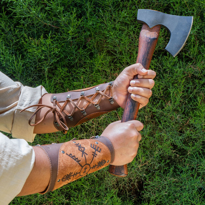 A model wearing Viking Bracers by Hide & Play while holding an axe, highlighting the impressive features of this set of 2 Viking Style Bracers. These arm protectors for costume are made from full grain leather, ideal for cosplay, featuring small batch production and artisanal craftsmanship from La Antigua Guatemala & Oaxaca Mexico.