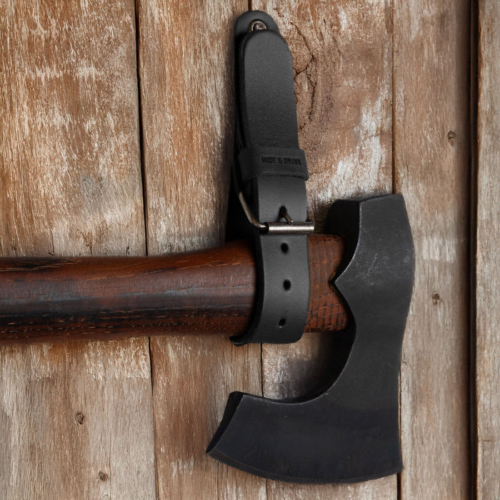 Close-up image of a black Hide & Drink Wall Hanging Strap holding an axe against wood, highlighting its sturdy and reliable craftsmanship. Featuring a buckle closure, this heavy-duty tool hanger adds style and organization, making it great for axes, hatchets & tools. Made from full grain leather, it is handmade, showcasing artisan quality from La Antigua Guatemala & Oaxaca Mexico.