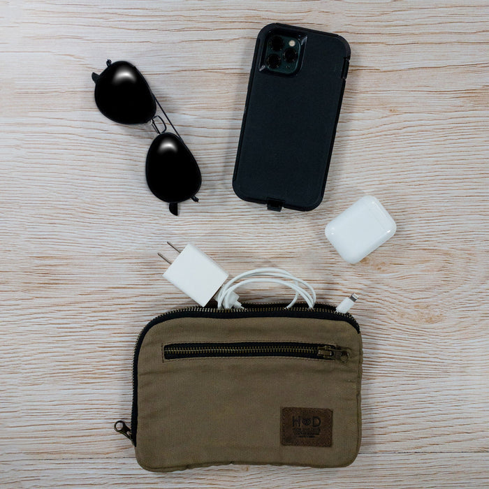 Hide & Drink's Zipper Gear Pouch alongside sunglasses and tech accessories, showcasing its utility as a travel bag crafted from water resistant waxed canvas.