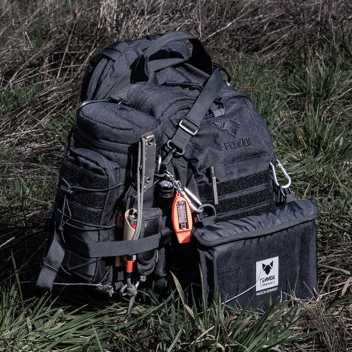 Der Backpack One2explore steht auf einer Wiese. Die Zubehörtasche ist seitlich mit den Kompressionsgurten am Backpack festgeschnallt. An der Vorderseite befindet sich der  Klapprost Titan in seiner dazugehörigen Tasche, mithilfe von Karabinern ist dieser am Backpack befestigt.